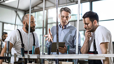 Deux jeunes employés de l'usine regardent des évaluations numériques d'une machine sur tablette avec leur collègue plus âgé dans un hall de production et en tirent des conclusions précieuses pour leur production.