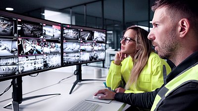 Photo : Deux personnes sont assises devant plusieurs écrans affichant des flux vidéo en direct et surveillent un système de sécurité et de vidéosurveillance.