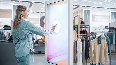 Une jeune femme blonde portant une veste en jean appuie sur le panneau de commande "Press Start" d'un grand écran d'information et de publicité pour une expérience de shopping moderne dans un magasin de mode.