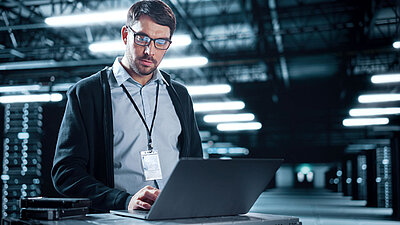 Un jeune collaborateur informatique avec des lunettes, une barbe de trois jours, une chemise bleu clair, un cardigan bleu foncé et un badge de sécurité autour du cou se tient dans une immense salle de serveurs grise et bleue et vérifie les paramètres de sécurité sur son ordinateur portable