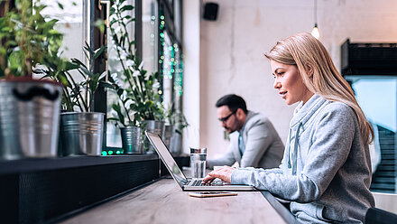 Photo de deux personnes travaillant sur leur ordinateur portable dans un bureau ou un café moderne ; à gauche, on voit de nombreuses plantes en pot sur le rebord de la fenêtre ; à droite, une jeune femme blonde au premier plan et un jeune homme brun à lunettes à l'arrière-plan sont assis devant leur ordinateur portable, face à la fenêtre, et travaillent sur quelque chose dans la bonne humeur.