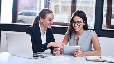 Une fonctionnaire avec une tresse et un blazer est assise à son bureau avec une collègue et lui montre comment remplir un formulaire numérique sur une tablette.