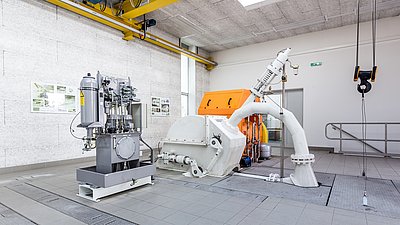 Installation de turbine industrielle avec système hydraulique dans une salle technique lumineuse, crochet de grue visible.