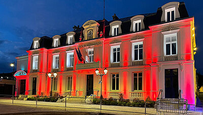 Hôtel de ville historique de nuit, illuminé en rouge et blanc, avec des drapeaux français et des lampadaires au premier plan