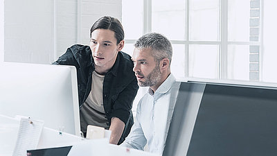Un jeune informaticien avec une tresse conseille un collègue d'âge moyen avec des cheveux gris et une barbe sur les paramètres de sécurité de l'ordinateur.