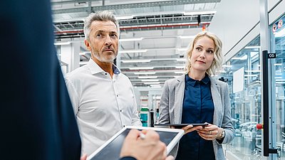 Photo : Deux personnes avec des tablettes dans un environnement industriel ou de production moderne avec des zones de machines vitrées et des installations techniques.