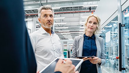 Photo : Deux personnes avec des tablettes dans un environnement industriel ou de production moderne avec des zones de machines vitrées et des installations techniques.