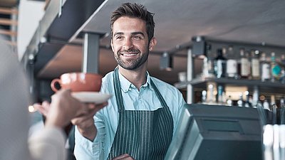 Un sympathique barista à la caisse, avec barbe et tablier, tend une tasse de café à une cliente en souriant