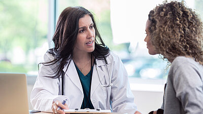 Un jeune médecin aux longs cheveux bruns et à la blouse médicale explique quelque chose à une patiente aux boucles brun clair à la table de consultation du cabinet médical