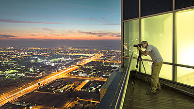 Photo d'un homme prenant une photo de la ville illuminée la nuit avec son trépied d'appareil photo depuis le balcon / toit de son appartement penthouse au-dessus d'une grande ville