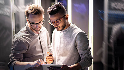 Deux hommes se trouvent dans une salle de serveurs et analysent ensemble des données sur une tablette.