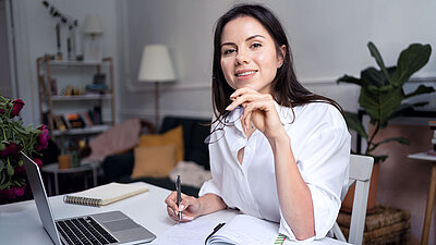 Jeune femme brune portant un chemisier d'affaires blanc, souriant avec satisfaction à la caméra et tenant ses lunettes qu'elle vient d'enlever, assise à son bureau dans son bureau à domicile, prenant des notes à côté de son ordinateur portable ; en arrière-plan, on voit des plantes, un lampadaire et une étagère de livres