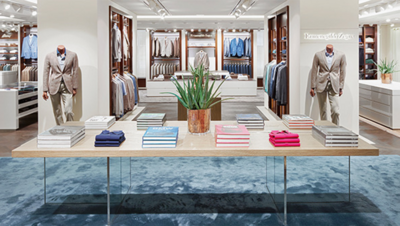 Photographie de la surface de vente du magasin de mode Lodenfrey : table de marchandises avec des chemises d'hommes élégamment arrangées dans différentes couleurs et une plante au premier plan et des étagères et des murs d'exposition bien assortis, y compris deux mannequins de mode masculine équipés de costumes d'hommes beiges à l'arrière-plan.