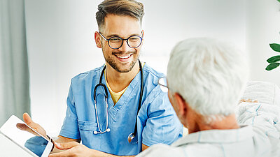 Photographie d'un jeune infirmier pour personnes âgées, blond et souriant, portant des lunettes, une blouse de médecin bleue et un stéthoscope autour du cou, montrant seulement un dossier à une dame âgée aux cheveux blancs courts, que l'on voit de dos au premier plan