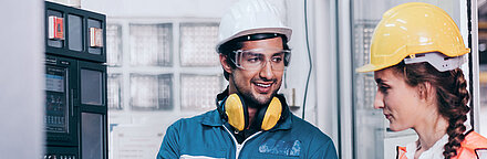 Un ouvrier aux cheveux noirs, portant un casque de protection blanc, des lunettes de protection et des écouteurs antibruit jaunes, vêtu d'une combinaison bleue et de gants de travail, tient un ordinateur portable à la main et se tient à droite d'un boîtier de commande (à gauche) ; il explique quelque chose à sa collègue brune, à sa droite, en souriant ; sa collègue porte également un casque jaune et un gilet de sécurité et écoute avec attention