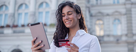 Une jeune femme aux cheveux bouclés et portant un chemisier se tient devant un bâtiment administratif et exécute avec satisfaction un processus administratif numérisé sur sa tablette, sa carte bancaire à la main.