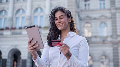 Une jeune femme aux cheveux bouclés et portant un chemisier se tient devant un bâtiment administratif et exécute avec satisfaction un processus administratif numérisé sur sa tablette, sa carte bancaire à la main.