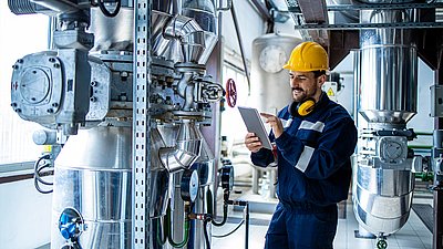 Photo : Technicien portant un casque de sécurité et une tablette au milieu de conduites industrielles, de vannes et de composants techniques d’installation.