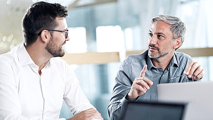 Vue de face de deux hommes assis à une table de conférence dans un bureau moderne et lumineux, en train de discuter avec animation devant un ordinateur portable : L'homme de gauche est trentenaire, brun, porte une barbe, des lunettes et une chemise blanche et regarde son collègue (à sa droite) avec enthousiasme ; l'homme de droite est d'âge moyen, porte des cheveux gris, une courte barbe grise et une chemise bleu pigeon à petits points blancs et explique quelque chose d'important à son collègue en pointant l'index droit vers le haut