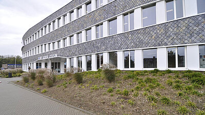 Photo du bâtiment scolaire de la Scholengroep Pontes : bâtiment moderne et incurvé avec de la pierre grise et de nombreuses fenêtres blanches, y compris une pelouse devant.