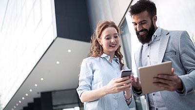 Photo : Deux personnes se tiennent devant un bâtiment moderne et regardent ensemble un smartphone et une tablette.