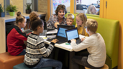 Photo d'une enseignante d'âge moyen aux cheveux frisés, travaillant et enseignant avec des élèves de primaire, hommes et femmes, dans un coin coloré équipé d'ordinateurs portables.
