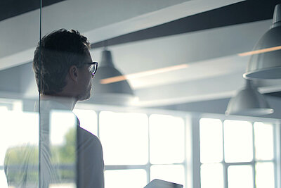 Un homme se tient derrière la paroi vitrée d'un bureau, une tablette à la main.