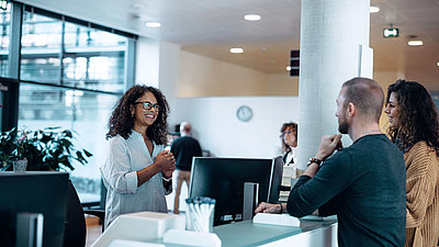 Une employée administrative aux cheveux noirs et aux lunettes accueille aimablement un jeune couple à son poste de travail numérique
