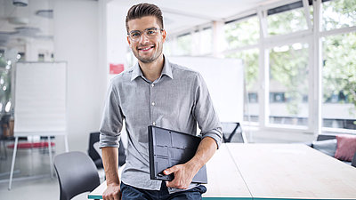 Un jeune fonctionnaire, lunettes et tablette à la main, se tient de manière souveraine dans un local administratif clair et moderne.
