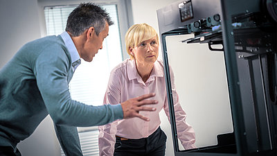 Une femme blonde d'âge moyen aux cheveux courts discute avec son collègue masculin devant une machine.