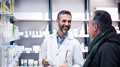 Deux hommes dans une pharmacie ; pharmacien aux cheveux noirs courts et à la barbe ; porte une blouse de médecin et tient un médicament à la main. Il sourit à un client qui se tient devant lui, également cheveux noirs et courts, veste et écharpe.