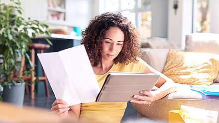 Femme assise devant le canapé, tenant une tablette dans la main gauche et un papier dans la main droite