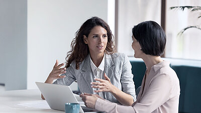 Secure Edge Computing Zwei Kolleginnen sitzen am Tisch vor einem Laptop, eine gestikuliert mit ihren Händen