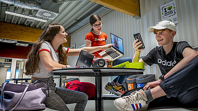 Photo prise dans les salles communes de l'Udens College : trois adolescents sont assis à une table avec leurs cartables et utilisent avec enthousiasme le réseau WLAN rapide de l'école ; à droite, un garçon blond, casquette et téléphone portable à la main, est assis et sourit ; à gauche, deux filles brunes aux cheveux longs sont assises ; l'une d'elles montre à l'autre quelque chose sur son ordinateur portable et rit.