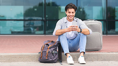 Un voyageur assis sur le trottoir devant le hall de l'aéroport avec son smartphone et sa valise
