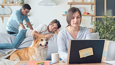 Une femme travaille à domicile sur son ordinateur portable avec sa fille, son chien et son mari en arrière-plan.